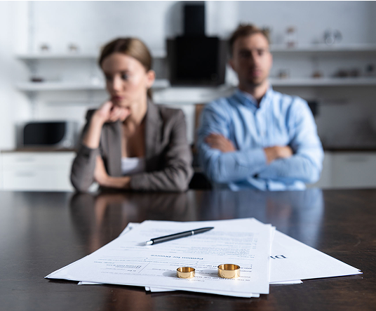 A couple sitting apart in a room, with their backs turned to each other. On a table in front of them, there are divorce papers and two gold wedding bands.