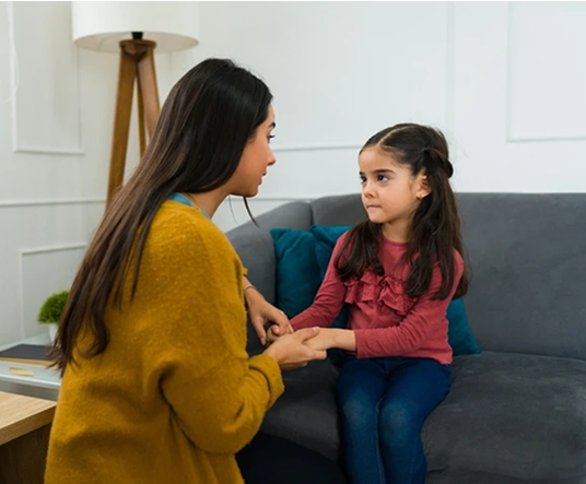 A woman sitting on a sofa, holding the hands of a young girl, who is looking at her with a serious expression. The room is brightly lit and features a modern decor.