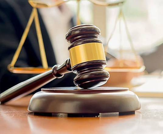 A wooden gavel resting on a circular base in the foreground, with a set of balance scales blurred in the background, suggesting a legal context.